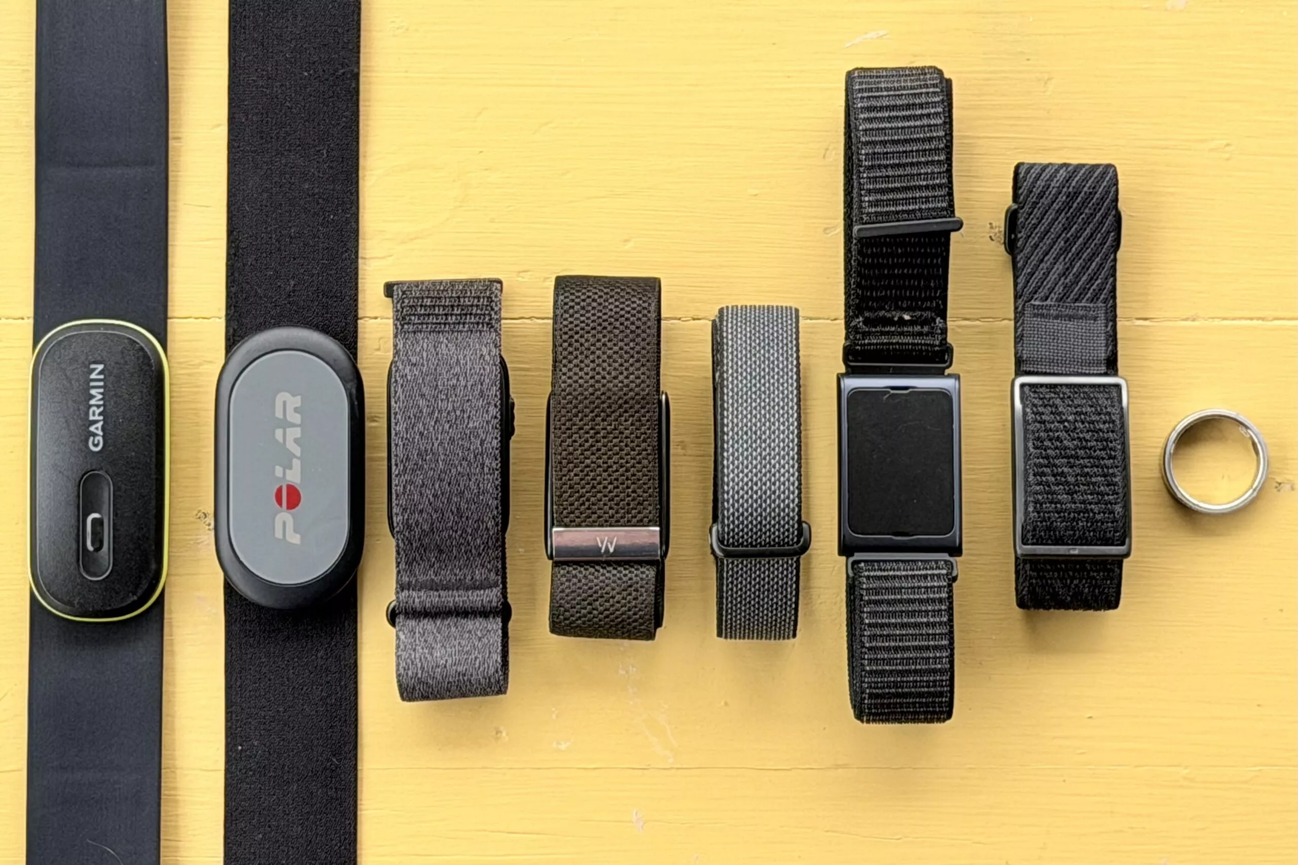 Perbandingan berbagai pelacak aktivitas (smart band dan smart ring) di permukaan kayu kuning, termasuk produk Garmin CIRQA tanpa layar.