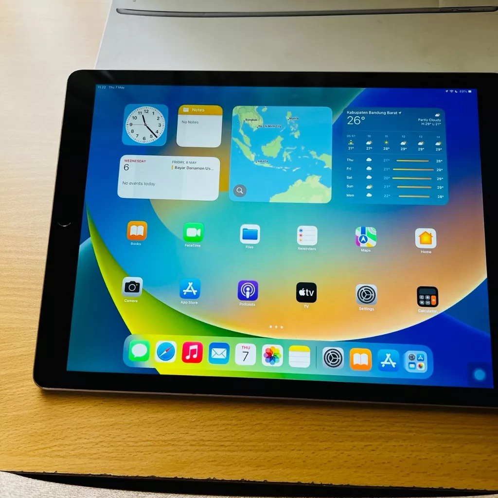 iPad Gen 12 diletakkan di atas meja dengan layar aktif yang menampilkan widget jam, peta, dan cuaca.