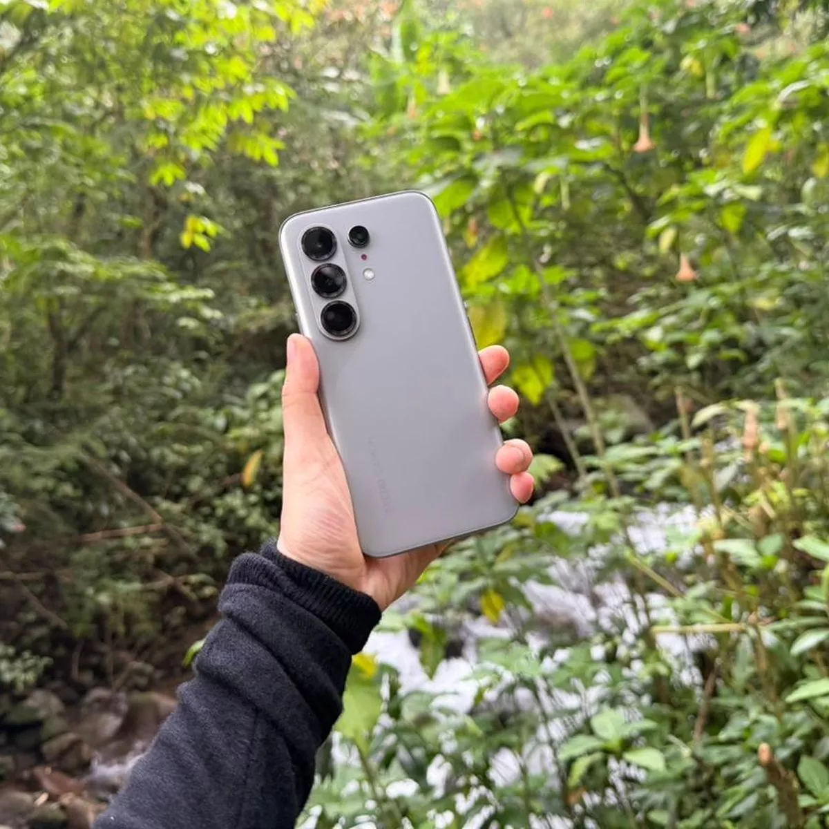 Smartphone 5G warna perak dipegang di luar ruangan dengan latar belakang pepohonan hijau.