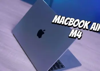 Tampilan fisik MacBook Air M4 berwarna abu-abu yang terbuka di atas meja kayu dengan pencahayaan biru.