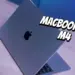Tampilan fisik MacBook Air M4 berwarna abu-abu yang terbuka di atas meja kayu dengan pencahayaan biru.