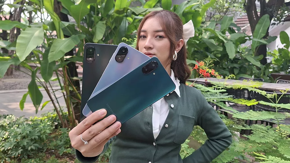 Wanita memegang tiga unit HP Low-Mid Range varian Samsung di luar ruangan dengan latar tanaman hijau.