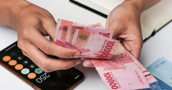 Tangan pekerja menghitung lembaran uang tunai rupiah BSU Kemenaker 2026 di dekat kalkulator ponsel