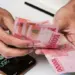 Tangan pekerja menghitung lembaran uang tunai rupiah BSU Kemenaker 2026 di dekat kalkulator ponsel